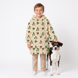A young boy stands next to a black and white dog wearing a beige hooded sweatshirt covered in a green cactus and cartoon animal print, paired with khaki pants and white socks, against a plain white studio background.