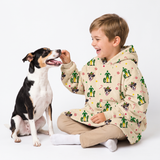 A young boy wearing a beige hoodie with a green and multicolor dog print sits cross-legged on a white studio floor, smiling as he offers a treat to a black, white, and tan dog.