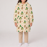 A child wearing a cream-colored hooded sweatshirt covered in a festive green, gold, and red elf pattern, paired with beige pants and white socks, standing against a light gray studio background.