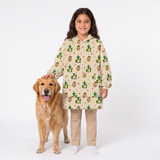 A young girl stands next to a golden retriever dog wearing a cream-colored hooded sweatshirt covered in a green cactus and sun pattern, paired with khaki pants and white socks, against a plain white studio background.