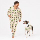 A man and a dog wearing matching cactus-print pajamas pose together against a plain white studio background. The pajamas feature a cream base with a green cactus and black cat pattern.