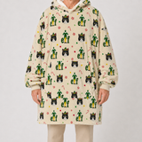 A person wearing a cream-colored hooded sweatshirt with an all-over festive print of green elf hats, black-and-white cat faces, candy canes, and small red and yellow paw prints, paired with light beige pants, against a plain light gray studio background.
