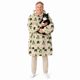 A man wearing a hooded beige robe with a black cat printed all over it, holding a black and white cat. He is also wearing beige pants and white socks, standing against a solid white background.