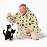 A man wearing a beige hooded blanket with a green, black, and red cat pattern sits on a white studio floor, smiling as a black and white cat reaches for a dangling toy.