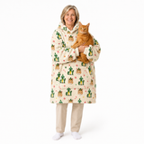 A smiling woman stands against a white studio background holding an orange tabby cat. She is wearing a hooded fleece pajama coat with a cream base and a repeating print of green cacti, small orange flowers, and orange cat faces.