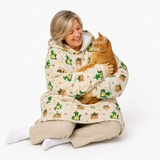 A woman sitting on a white studio floor holds an orange tabby cat wrapped in a cream-colored hooded blanket printed with green cacti and small cartoon faces.