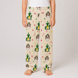 A child wearing beige holiday-themed lounge pants with a repeating print of green elf figures, red candy canes, paw prints, and dog faces, standing barefoot against a light gray studio background.