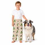 A young boy stands barefoot next to an Australian Shepherd dog, wearing a pair of beige pajama pants with a green cactus and dog print, paired with a plain white t-shirt, against a solid white background.