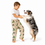 A young boy wearing beige Christmas-themed pajamas with a cactus and Santa pattern, standing barefoot and smiling as he plays with a black, white, and tan Australian Shepherd dog on a white studio background.
