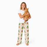 A young girl stands barefoot against a white studio background, holding an orange tabby cat. She is wearing a white short-sleeve T-shirt and cream pajama pants printed with cactus and candy cane motifs.