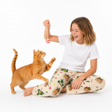 A child sitting on a white studio floor playing with a ginger cat, wearing a white t-shirt and cream pajama pants with a cartoon cat print. The child holds up a small red-and-white striped toy above the cat.