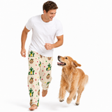 A man and a golden retriever dog wearing matching Christmas-themed pajamas, running on a solid white background. The pajama pants are cream-colored with a festive pattern of elves, candy canes, and snow globes.
