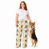 A woman wearing cream-colored pajama pants with a festive elf and corgi print stands next to a corgi dog on a white studio background. The pajama pants feature green elf hats, brown corgi faces, and red candy cane details.