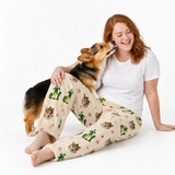 A woman with red hair sits on a white studio floor wearing a white t-shirt and beige Christmas-themed pajama pants with a corgi dog on her lap.