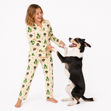 A woman wearing a cream-colored pajama set with a green cactus and black cat pattern is playfully interacting with a black and white dog on a plain white background.