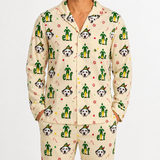 A person wearing a matching long-sleeve pajama top and pants set in a warm cream color, covered in a festive Christmas elf print featuring Dalmatian dogs, green hats, candy canes, and paw prints.