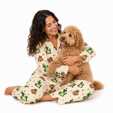 A woman and her golden doodoodle dog are sitting together on a white studio background, both wearing matching cream-colored pajamas with a festive green, red, and brown holiday print featuring Santa Claus, reindeer, and paw prints.