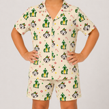 A child wearing a matching short-sleeve pajama shirt and shorts set in cream, covered in a festive print of Dalmatian dogs dressed as elves, candy canes, and snowflake motifs, photographed against a light gray studio background.
