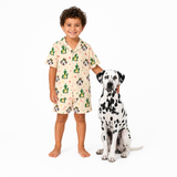 A young boy wearing a short-sleeve pajama shirt and matching shorts with a cute dog-themed print, standing next to a Dalmatian dog on a solid white background.