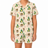 A child wearing a matching short-sleeve pajama set with a cream background and an all-over festive print featuring green elf silhouettes, fox faces, candy canes, and small holiday motifs.