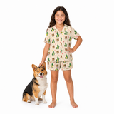 A young girl stands barefoot next to a corgi dog on a white studio background, wearing a matching short-sleeve pajama set with a cream base and a green, brown, and red illustrated print.