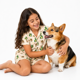 A young girl with long dark hair sits on a white studio floor smiling at a corgi dog. She is wearing a short-sleeve pajama set with a beige background and a green cactus and animal print.