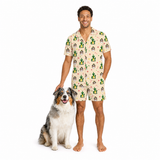 A man wearing a matching beige short-sleeve pajama set with a green and pink dog print stands next to an Australian Shepherd dog on a white background.