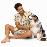 A man wearing a beige short-sleeve pajama shirt and matching shorts with a green cactus and dog print sits on a white studio floor, smiling as he brushes a black, white, and tan Australian Shepherd. A brown leather leash lies on the floor nearby.