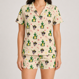A woman modeling a matching short-sleeve button-front pajama top and shorts set in a warm cream color, covered in a festive Christmas elf pattern with green elves, Santa hats, candy canes, and dog faces.