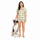 A woman wearing a matching short-sleeve pajama set with a dog printed pattern stands next to a black and white dog on a solid white background. The pajama set has a beige base with green, brown, and black cactus and animal motifs.