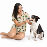 A woman wearing a beige short-sleeve pajama set with a green cactus print sits on the floor next to a black and white dog, offering a small treat. The background is a clean white studio backdrop.