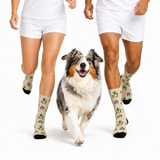A dog and two cropped human legs wearing matching beige socks with a green and gray dog-themed pattern, all on a clean white studio background.
