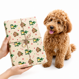 A brown curly-haired dog sits next to a gift box wrapped in beige holiday-themed wrapping paper with cartoon characters, held by hands against a white background.