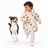 A child running happily alongside a black, white, and tan dog on a seamless white studio background. The child is wearing a cream-colored hooded sweatshirt covered in a colorful retro pattern of hot air balloons, rockets, and rockets launching, paired with beige pants and white socks.