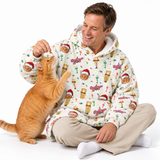 A man wearing a white fleece hoodie with a Christmas-themed cat print sits cross-legged on a white studio floor, smiling as an orange tabby cat jumps up to lick his face. The hoodie features a repeating pattern of cats in Santa hats, Christmas trees, and ornaments, with 'Ojibba' text printed on the sleeves.