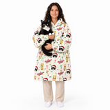 A woman wearing a cream-colored hooded robe with a festive Christmas print of Santa Claus sleighs, misty mountains, and snow globes, holding a black and white cat. She is standing against a solid white background.