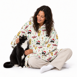 A woman sitting on a white studio floor wearing a hooded fleece pajama set with a festive holiday print of cats in Santa hats and candy canes, next to a black and white cat.