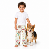 A young boy wearing a white t-shirt and Christmas-themed dog-print pajama pants stands next to a corgi dog on a seamless white background.