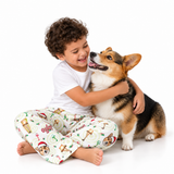 A young boy with curly brown hair sits cross-legged on a white studio floor, hugging a corgi dog. He is wearing a white t-shirt and cream-colored pajama pants printed with Christmas-themed illustrations including a Santa Claus dog, candy canes, and a snowman.