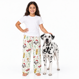 A young girl wearing a white t-shirt and Christmas-themed pajama pants stands next to a Dalmatian dog on a clean white background. The pajama pants feature a festive pattern of Santa-clad Dalmatians, candy canes, and holiday decorations.