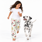 A young girl and a Dalmatian dog running and jumping on a white studio background. The girl is wearing a white t-shirt and cream-colored pajama pants with a Christmas print featuring Dalmatian dog faces, Santa hats, and holiday motifs.