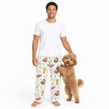 A man wearing a white t-shirt and patterned pajama pants stands barefoot next to a small brown dog on a plain white studio background. The pajama pants have an off-white base with a festive print of Santa-clad dogs, hot dogs, and holiday decorations.