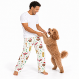 A man and his brown poodle dog play together while wearing matching white holiday-themed pajama pants with a festive print of Santa clowns, Christmas trees, and snow globes.