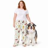 A woman wearing white pajama pants with a Christmas-themed dog print stands next to an Australian Shepherd dog on a white studio background. The pajama pants feature a cream base with a pattern of dogs in Santa hats, candy canes, and holiday decorations.