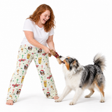 A woman with red hair wearing a white t-shirt and Christmas-themed dog-print pajama pants is playfully tug-of-war with a black, white, and tan dog on a white studio background.