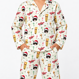 A person wearing a matching long-sleeve pajama top and pants set with an all-over festive print. The pajamas feature a cream background with repeated illustrations of cats in Santa hats, musical instruments, cocktail glasses, and red script text, photographed against a plain light gray studio background.
