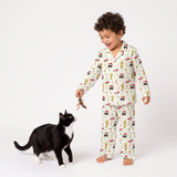 A young child wearing a long-sleeve pajama set with a festive Santa Claus and reindeer print, interacting playfully with a black and white cat on a clean white studio background.