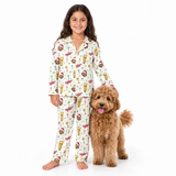 A young girl stands barefoot on a white studio background wearing a long-sleeve pajama set with an all-over food-themed print, next to a fluffy brown dog. The pajamas have a cream base with illustrations of burgers, fries, pizza, drinks, and holiday motifs.