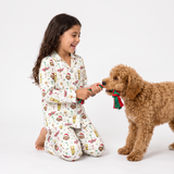 A young girl with long dark hair kneels on a white studio floor, smiling as she plays with a brown curly-haired dog. Both are wearing matching holiday-themed pajamas with a white background and a colorful print of Christmas treats, ornaments, and snowman motifs.