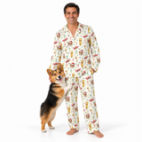 A man wearing a matching long-sleeve pajama set with a corgi standing beside him. The pajamas are off-white with a festive holiday print featuring Santa Claus, reindeer, snowflakes, and Christmas trees.